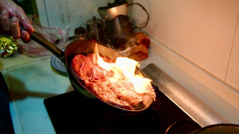 Close-up of a steak being seared with flames on a stovetop in a kitchen.