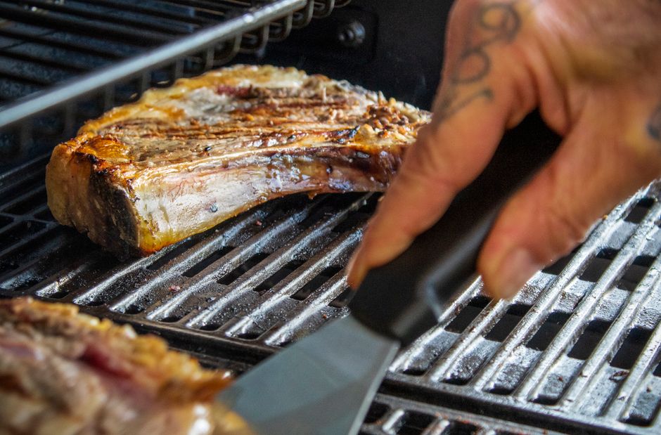Close-up of a ribeye steak being grilled on a barbecue with a spatula.